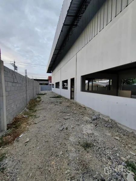Factory for rent or sale in Mueang Samut Prakan, Samut Prakan - Narrow service lane along the factory wall with windows and a perimeter block fence.