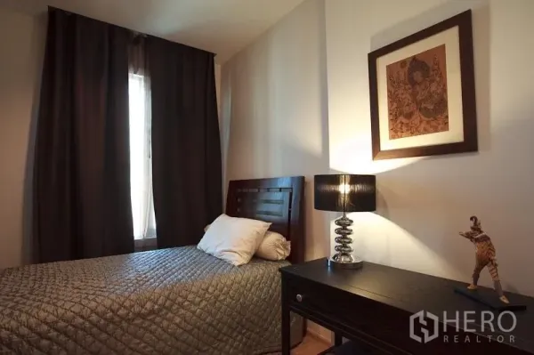 Condominium for rent in Khlong Toei, Bangkok - Cozy secondary bedroom with single bed, bedside lamp and study desk by a window.