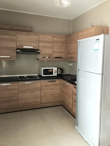 Condominium for rent in Watthana, Bangkok - L-shaped kitchen with wood cabinetry, cooktop, hood, microwave, and full-size fridge.