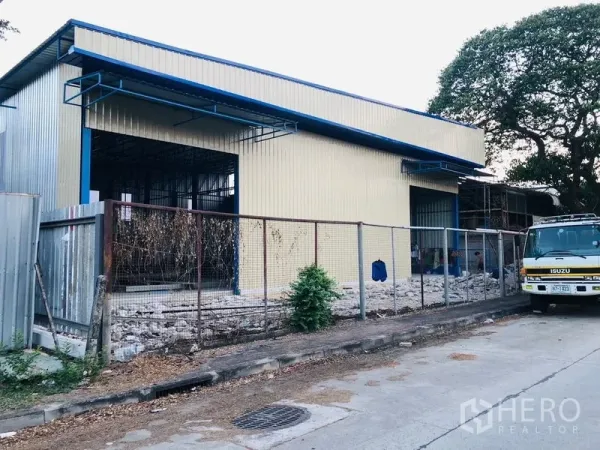 Warehouse for rent in Wang Thonglang, Bangkok - Street view of a yellow metal warehouse with blue roof, fenced frontage and a wide loading bay opening.