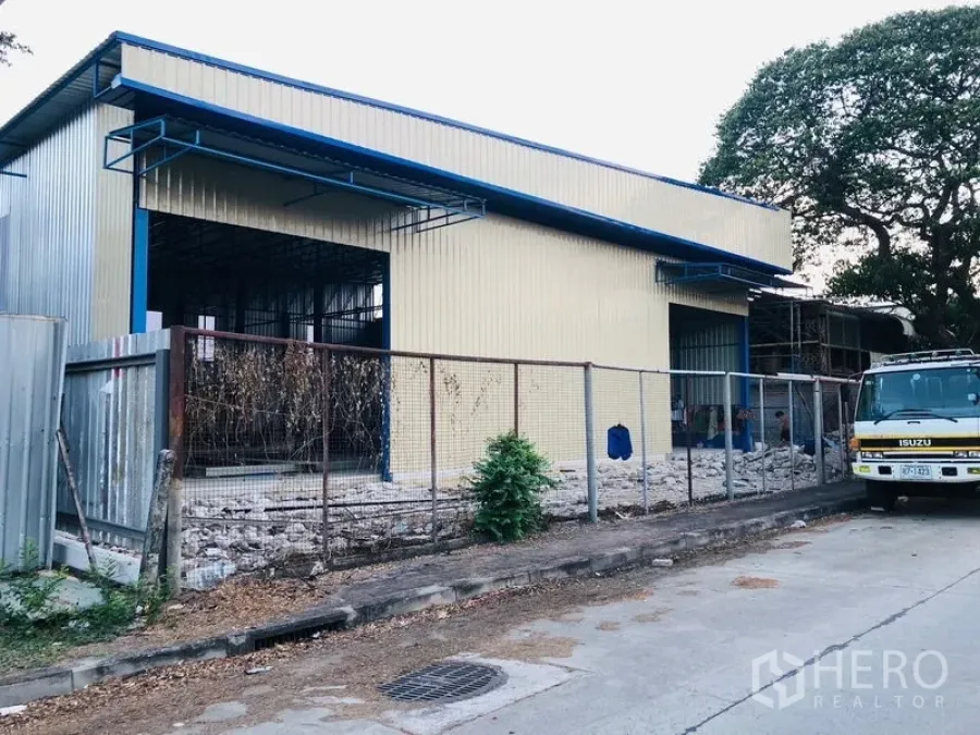 Warehouse for rent in Wang Thonglang, Bangkok - Street view of a yellow metal warehouse with blue roof, fenced frontage and a wide loading bay opening.