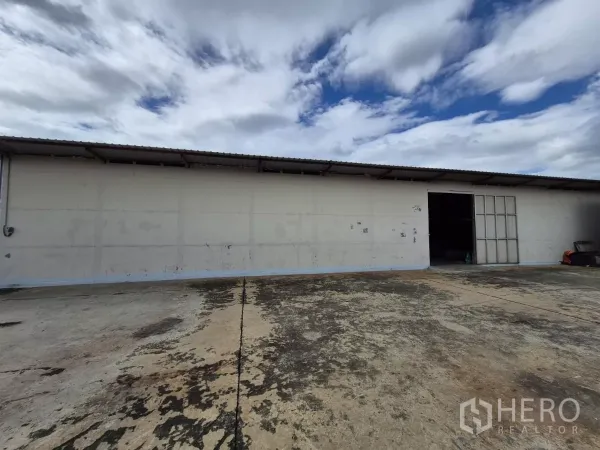 货栈 出租在Mueang Pathum Thani, Pathum Thani - 单层仓库外立面，金属推拉门，前方宽阔混凝土装卸场地，天空多云。