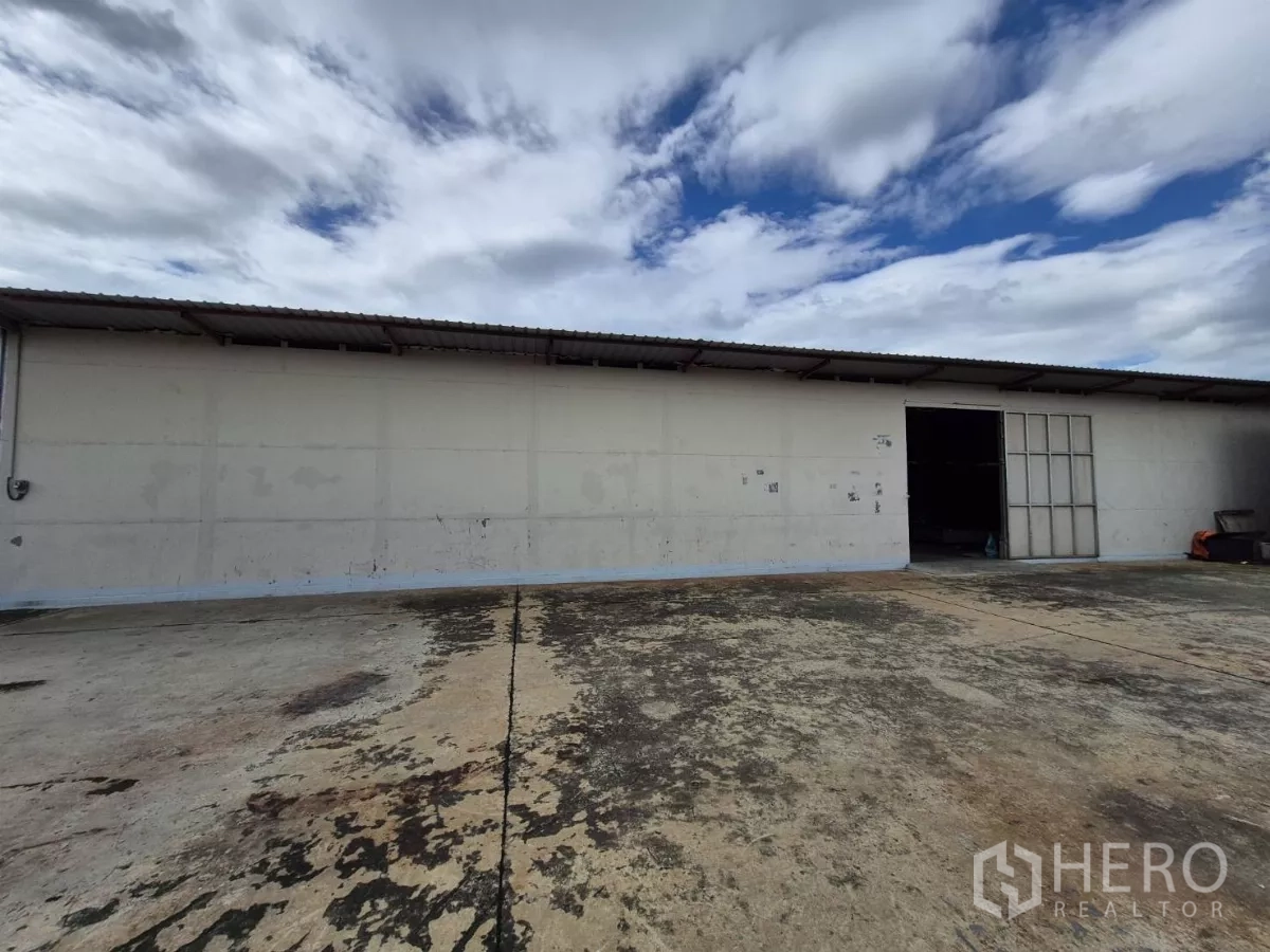 货栈 出租在Mueang Pathum Thani, Pathum Thani - 单层仓库外立面，金属推拉门，前方宽阔混凝土装卸场地，天空多云。