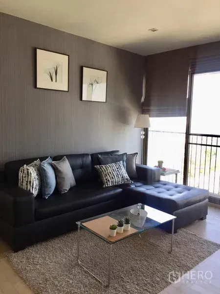 Condominium for rent in Watthana, Bangkok - Bright lounge featuring black sectional sofa with cushions, glass coffee table on rug, floor lamp, and balcony railing outside.
