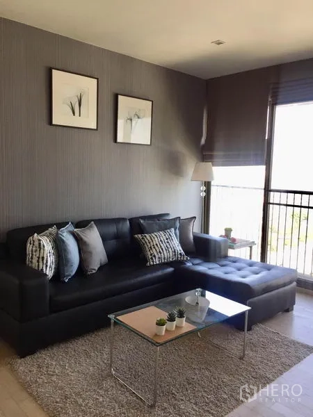 Condominium for rent in Watthana, Bangkok - Bright lounge featuring black sectional sofa with cushions, glass coffee table on rug, floor lamp, and balcony railing outside.
