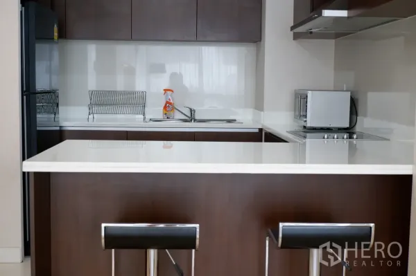 คอนโด ให้เช่าในPathum Wan, Bangkok - ครัวยุโรปท็อปหินสีขาว ตู้สีน้ำตาล เตาไฟฟ้า ไมโครเวฟ อ่างล้างจาน และเคาน์เตอร์บาร์พร้อมสตูล