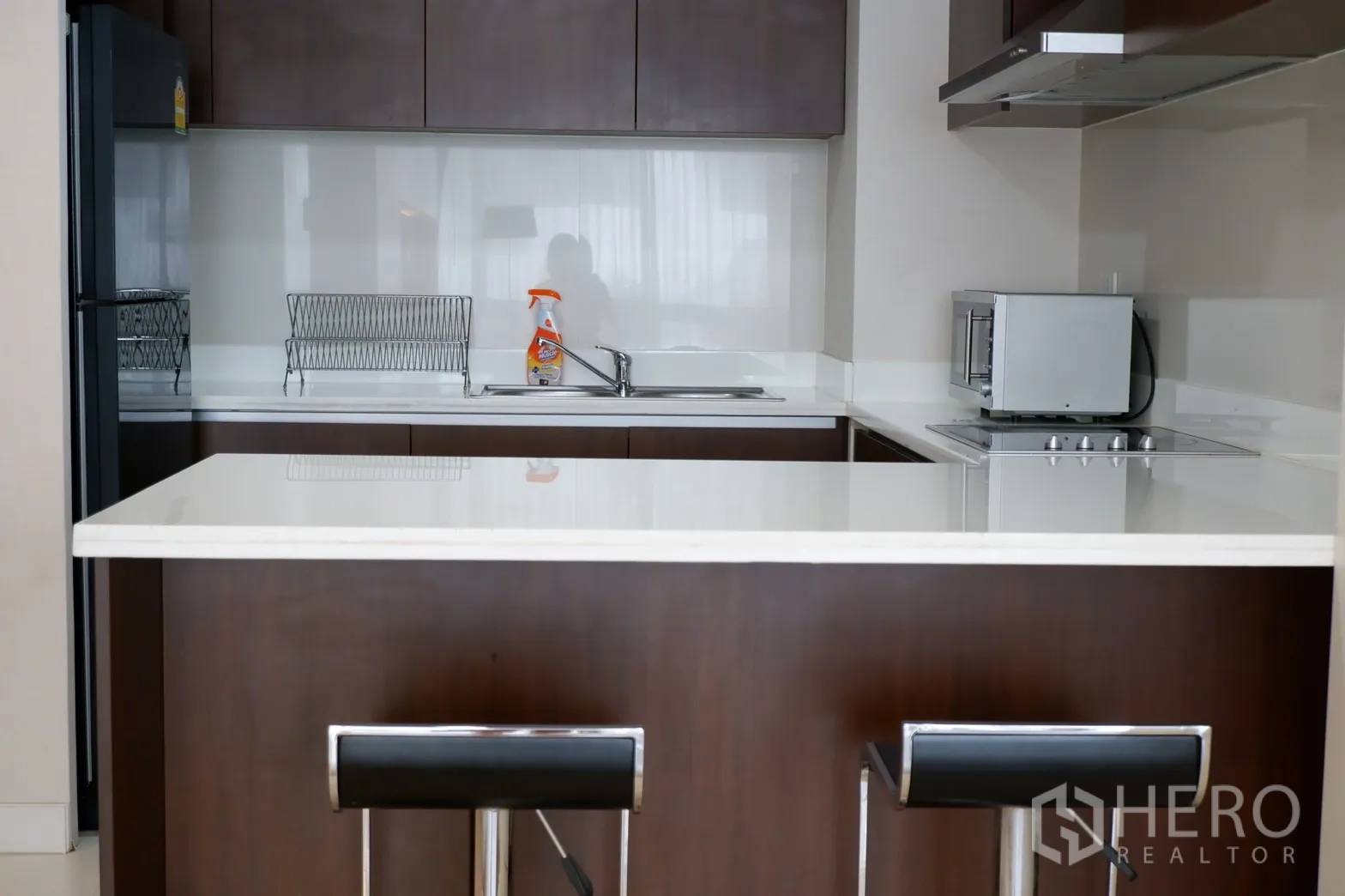 คอนโด ให้เช่าในPathum Wan, Bangkok - ครัวยุโรปท็อปหินสีขาว ตู้สีน้ำตาล เตาไฟฟ้า ไมโครเวฟ อ่างล้างจาน และเคาน์เตอร์บาร์พร้อมสตูล