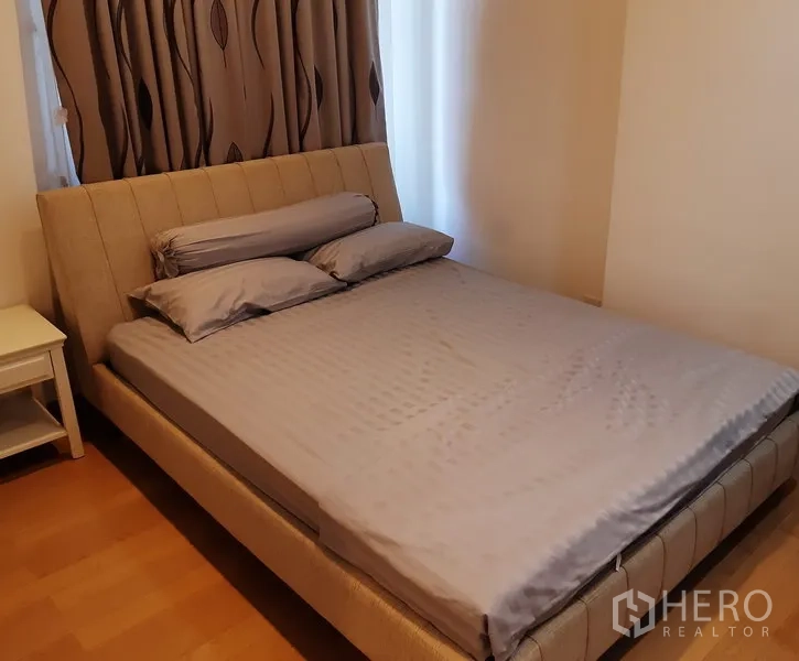Condominium for rent in Khlong Toei, Bangkok - Cozy bedroom with upholstered bedframe, bedside table and patterned blackout curtains in a 1-bedroom unit.