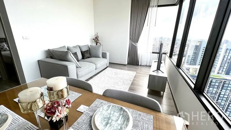 Condominium for rent in Watthana, Bangkok - Dining table set for four facing a gray sofa and TV, with panoramic city views through large windows.