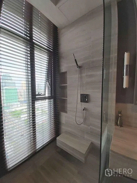 Condominium for sale in Bang Rak, Bangkok - Walk-in shower with built-in bench, gray tiles, and a floor-to-ceiling window with blinds overlooking the city.