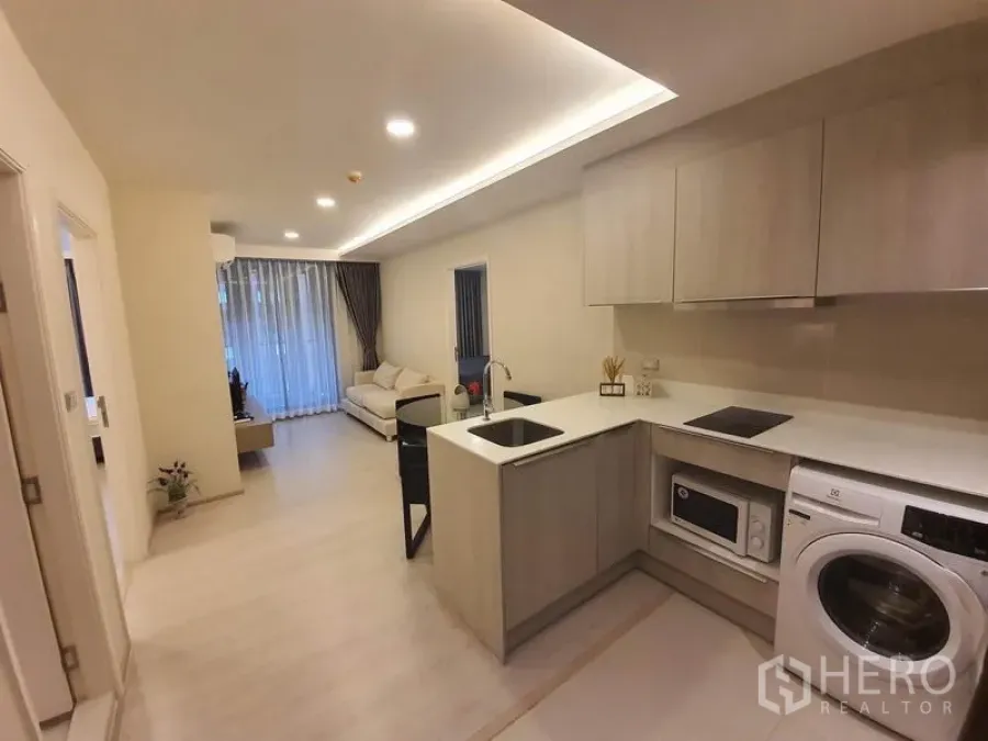 公寓 出租在Khlong Toei, Bangkok - 开放式厨房，带橱柜、洗衣机与中岛，连接明亮客厅与落地窗帘。