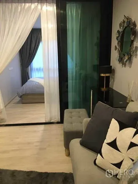 คอนโด ขายในBang Na, Bangkok - ห้องนั่งเล่นพร้อมโซฟาสีเทาและพัฟ มองเห็นฉากกั้นกระจกบานเลื่อนไปยังห้องนอนพร้อมผ้าม่านโปร่ง