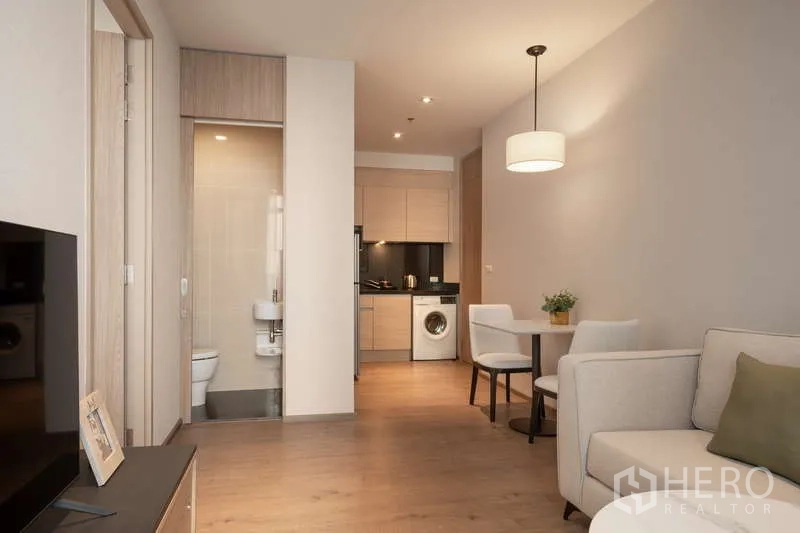 Condominium for rent in Khlong Toei, Bangkok - Open-plan living and dining area with round table, view toward a compact kitchen and bathroom entrance.