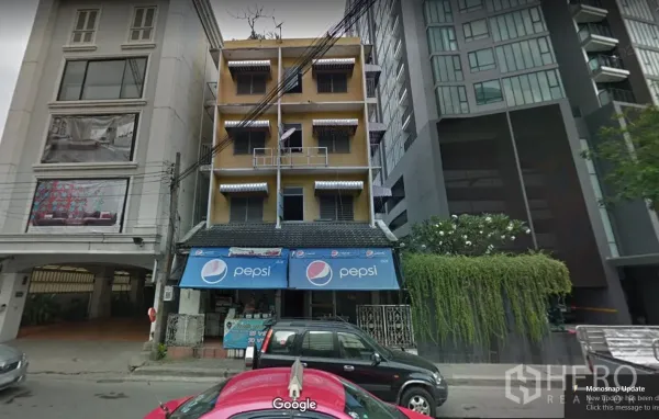 公寓 出租在Pathum Wan, Bangkok - 街景：带蓝色雨棚的临街店铺与现代高层公寓外立面相邻，周边有绿化和车流，靠近曼谷帕吞旺区的BTS拉差当利站。