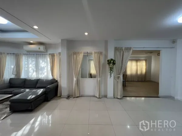 Detached House for rent in Bang Kapi, Bangkok - Lounge with L-shaped sofa, sheer curtains and doorway to a multi-purpose room.