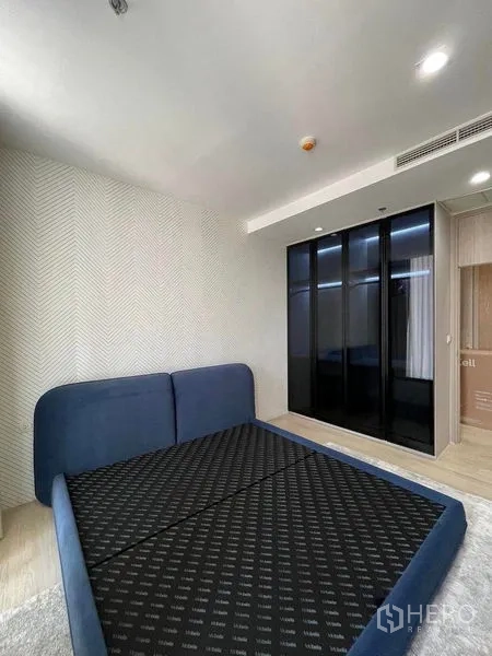 Condominium for rent in Pathum Wan, Bangkok - Minimalist bedroom with blue bed frame, herringbone wall and black glass wardrobe.