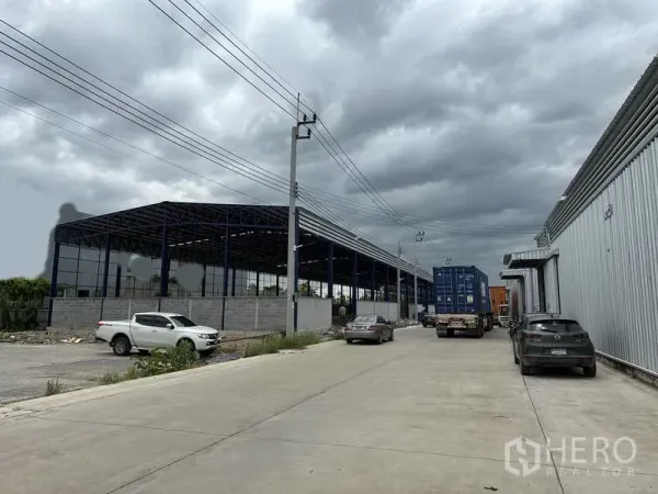 คลังสินค้า ให้เช่าในLat Krabang, Bangkok - มุมมองถนนอุตสาหกรรม มีรถคอนเทนเนอร์ใกล้โกดัง และโครงสร้างหลังคาเหล็กกำลังก่อสร้าง