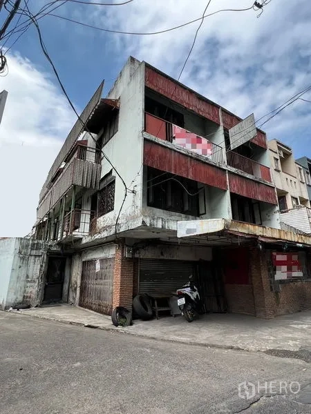 Shophouse for rent or sale in Bang Khen, Bangkok - Three-storey corner shophouse with rolling shutters and exposed brick at the ground level.