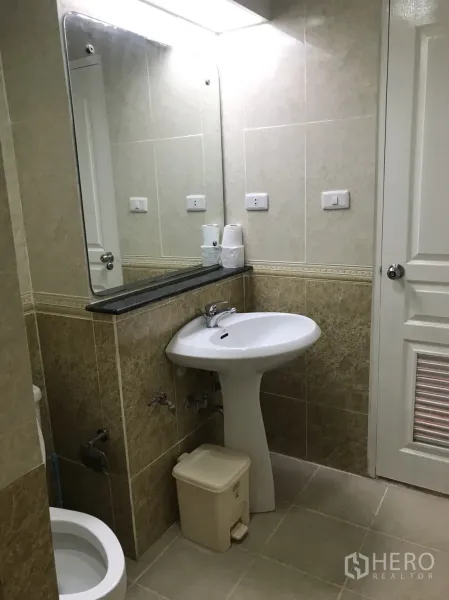 Condominium for rent in Khlong Toei, Bangkok - Tiled bathroom with pedestal sink, large mirror, and toilet in neutral tones.