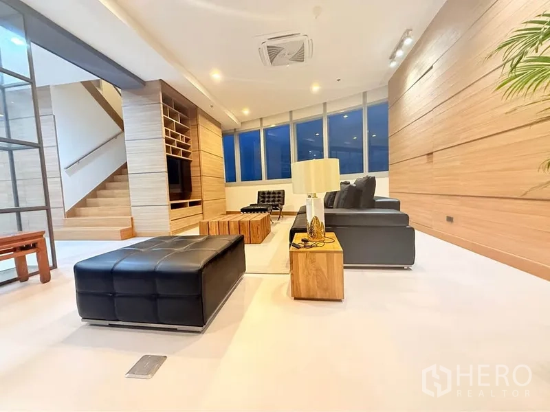 คอนโด ให้เช่าหรือขายในKhlong Toei, Bangkok - ห้องนั่งเล่นกว้าง โซฟาหนังสีดำ ผนังไม้ บิวท์อิน บันได และหน้าต่างบานใหญ่ยามค่ำ