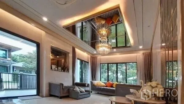 Detached House for rent or sale in Prawet, Bangkok - Double-height living room with chandelier, sofas, and floor-to-ceiling windows opening to a balcony.