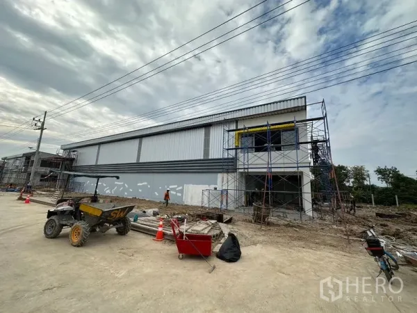 Warehouse for rent or sale in Lam Luk Ka, Pathum Thani - Side elevation of a new steel‑clad warehouse with scaffolding and tools along the roadside.