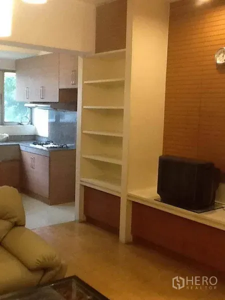 Condominium for rent in Pathum Wan, Bangkok - Open-plan living area leading to a windowed kitchen with wood cabinetry, gas cooktop, and built-in shelving.