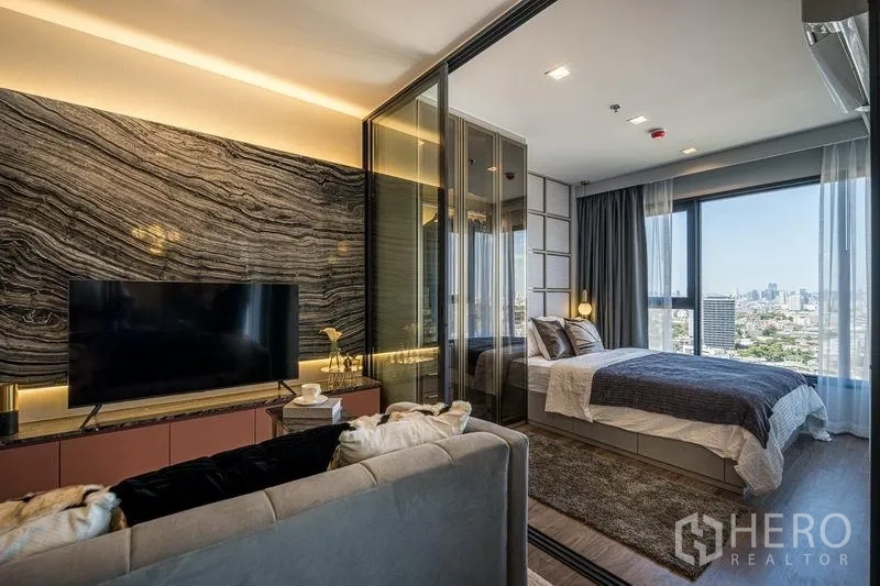 Condominium for sale in Chatuchak, Bangkok - Modern living room with stone-pattern feature wall, TV console, and glass-partitioned bedroom overlooking the Bangkok skyline.