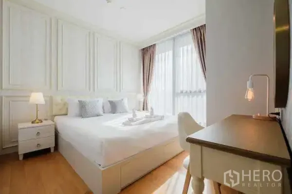 Condominium for rent in Khlong Toei, Bangkok - Bright bedroom with white panelled walls, king bed, writing desk and floor-to-ceiling window with curtains.