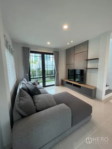 Detached House for rent or sale in Bang Phli, Samut Prakan - Bright living room with a grey sectional sofa, sliding glass doors, and a built-in TV wall.