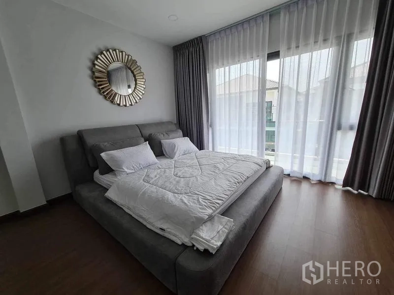 Detached House for rent or sale in Bang Phli, Samut Prakan - Cozy bedroom with grey upholstered bed, full-height windows and balcony view.