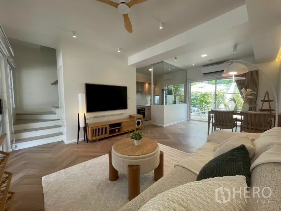 联排别墅 出租在Prawet, Bangkok - 明亮客厅，电视墙、用餐区、玻璃隔断厨房与花园景观。