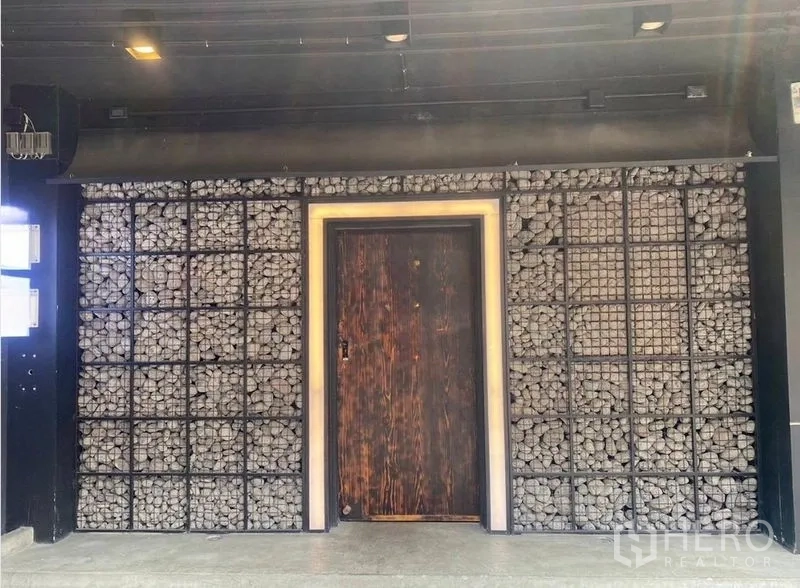 อาคารพาณิชย์ ให้เช่า - ด้านหน้าอาคารพาณิชย์ตกแต่งผนังตะกร้าหินและประตูไม้ใต้ไฟส่องสว่างสีเข้ม