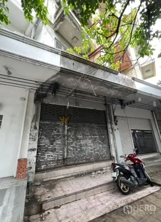 店屋 出租在Watthana, Bangkok - 临街店屋外立面，金属卷闸门，树荫下停放一辆摩托车。