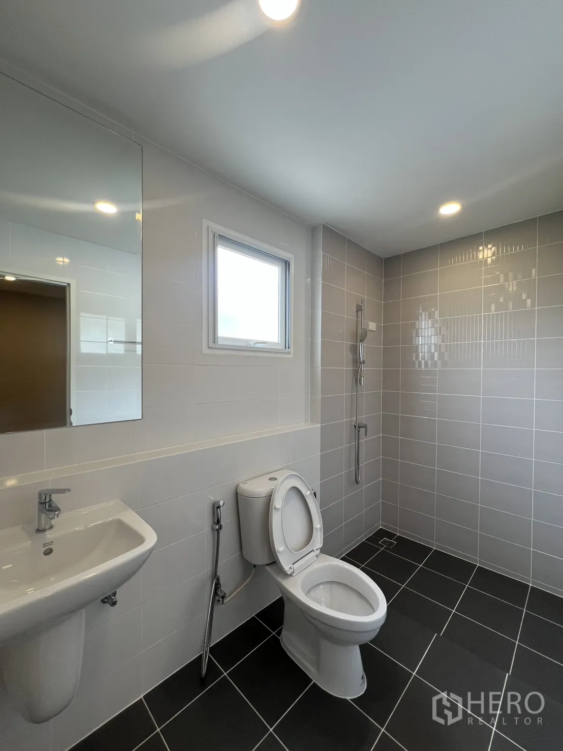 Detached House for rent in Lat Krabang, Bangkok - Modern bathroom with sink, toilet, handheld shower, and gray wall tiles with a small window.