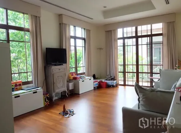 Detached House for rent in Watthana, Bangkok - Spacious family room with wood floors, tall windows, a TV console, and built-in toy storage.