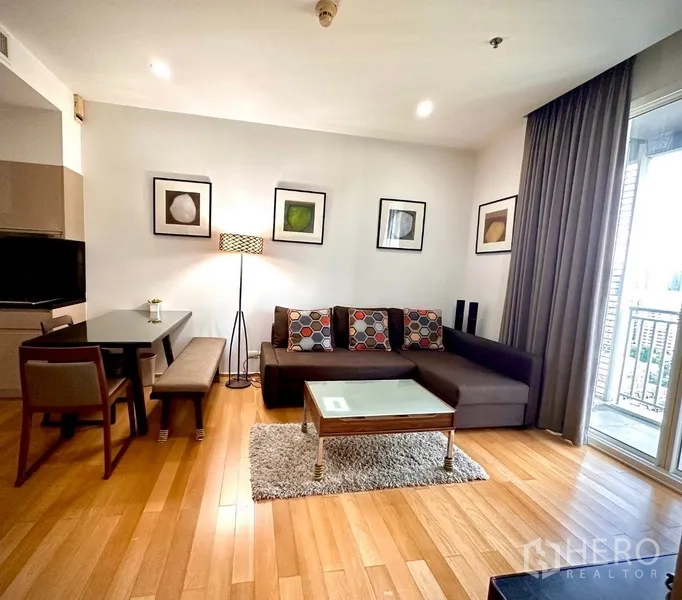 Condominium for rent or sale in Watthana, Bangkok - Living area with L-shaped sofa, area rug and balcony access offering partial city views.