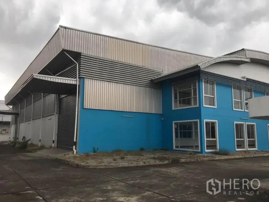 工厂 出租在Samut Prakan - 现代蓝色工厂外观，带两层办公楼、金属覆层与宽阔混凝土场地，天空多云。