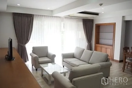 Condominium for rent in Pathum Wan, Bangkok - Living area with large windows, sheer curtains and a grey sofa set.