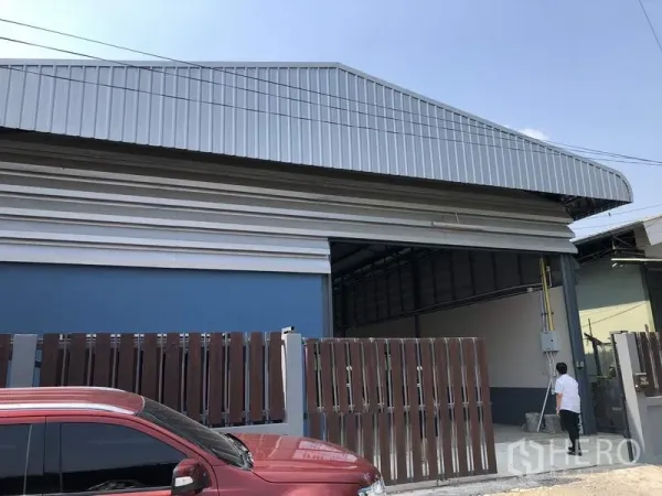 货栈 出售在Pathum Thani - 仓库外观，金属板墙与高山形屋顶，前方为棕色推拉围栏与车道。