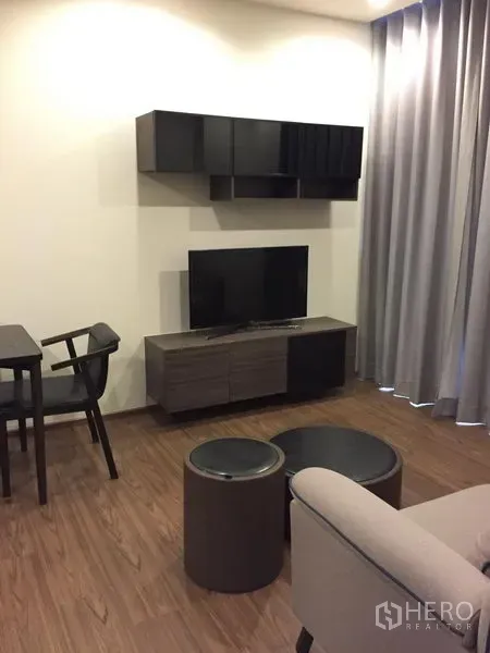 Condominium for rent in Watthana, Bangkok - Modern living room with wall-mounted shelves, TV console, small dining set, wood flooring and grey blackout curtains.