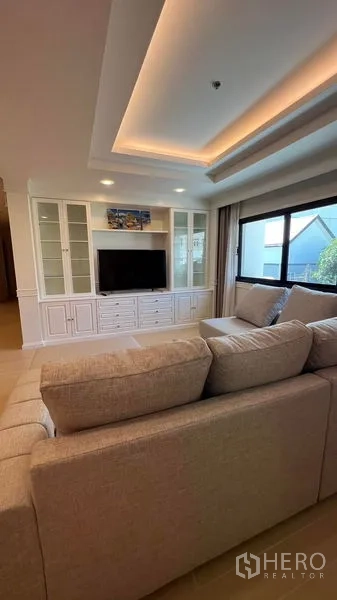 公寓 出租在Watthana, Bangkok - 客厅配L型沙发与电视柜展示墙，玻璃柜体及大窗户，可见绿意景观。