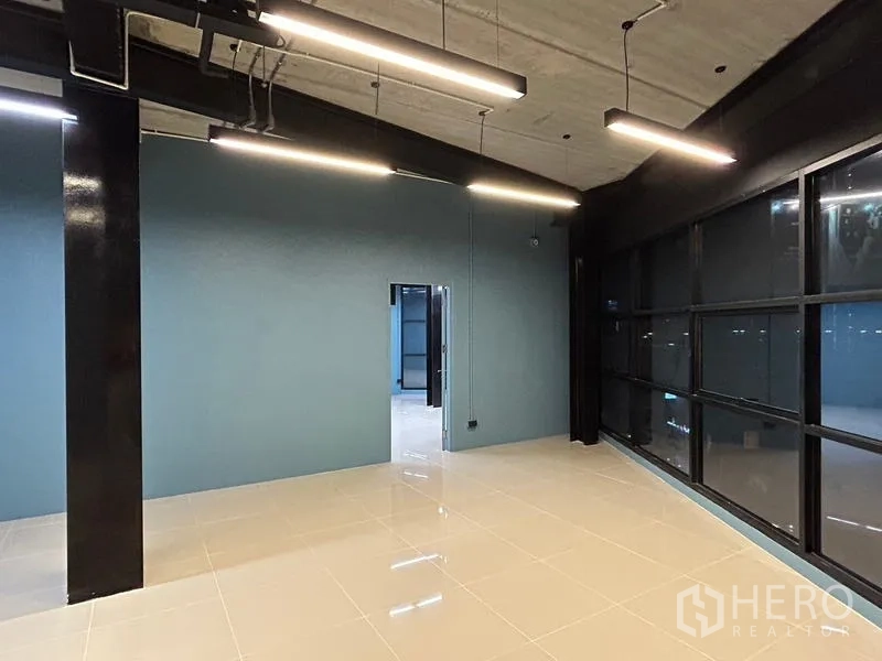办公空间 出租在Chatuchak, Bangkok - 开放式办公室，亮面瓷砖地面，青蓝色特色墙，外露天花与线性LED灯，旁有黑框窗。