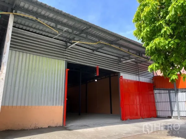 Warehouse for rent in Din Daeng, Bangkok - Front view of the warehouse under a tree with open red doors and high-clearance entry.