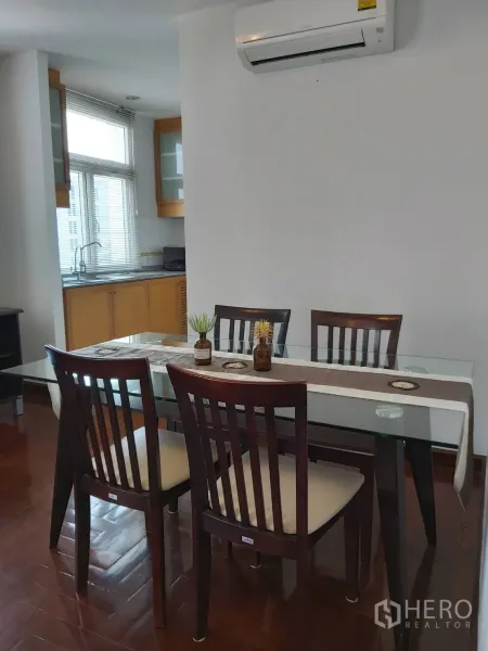 Apartment for rent in Khlong Toei, Bangkok - Glass-top dining table with six wooden chairs beneath a wall-mounted air conditioner next to the kitchen.