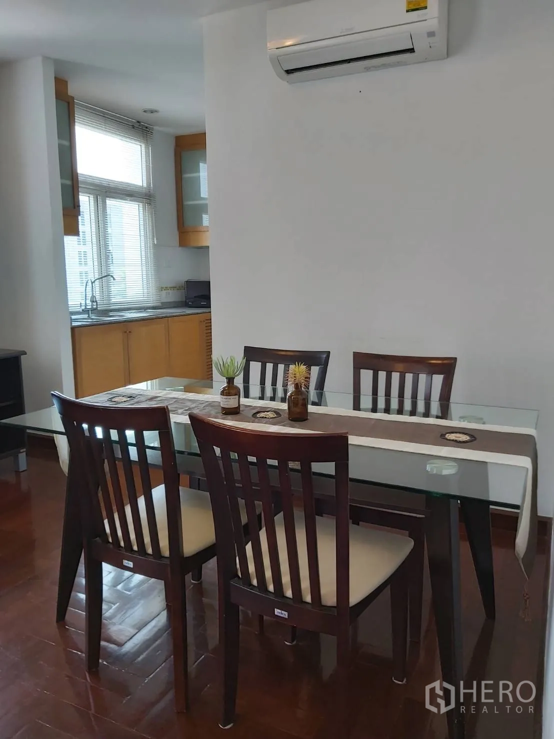 Apartment for rent in Khlong Toei, Bangkok - Glass-top dining table with six wooden chairs beneath a wall-mounted air conditioner next to the kitchen.