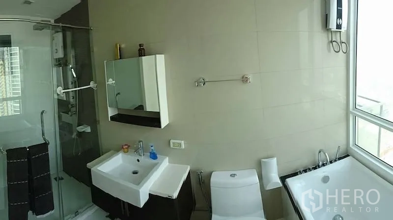 Condominium for rent or sale in Watthana, Bangkok - Modern bathroom with vanity, toilet, bathtub by a large window and a glass shower enclosure.