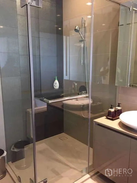 Condominium for rent in Bang Rak, Bangkok - Glass-enclosed shower with handheld spray next to vanity and mirrored cabinet.