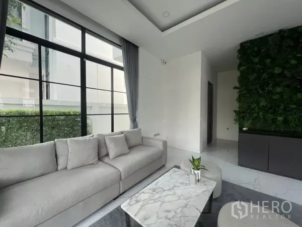 บ้าน ให้เช่าในLat Phrao, Bangkok - มุมนั่งเล่นร่วมสมัย โซฟาสีเทา โต๊ะกลางหินอ่อน และหน้าต่างกรอบอลูมิเนียมสูงชิดเพดานข้างผนังสวนแนวตั้ง