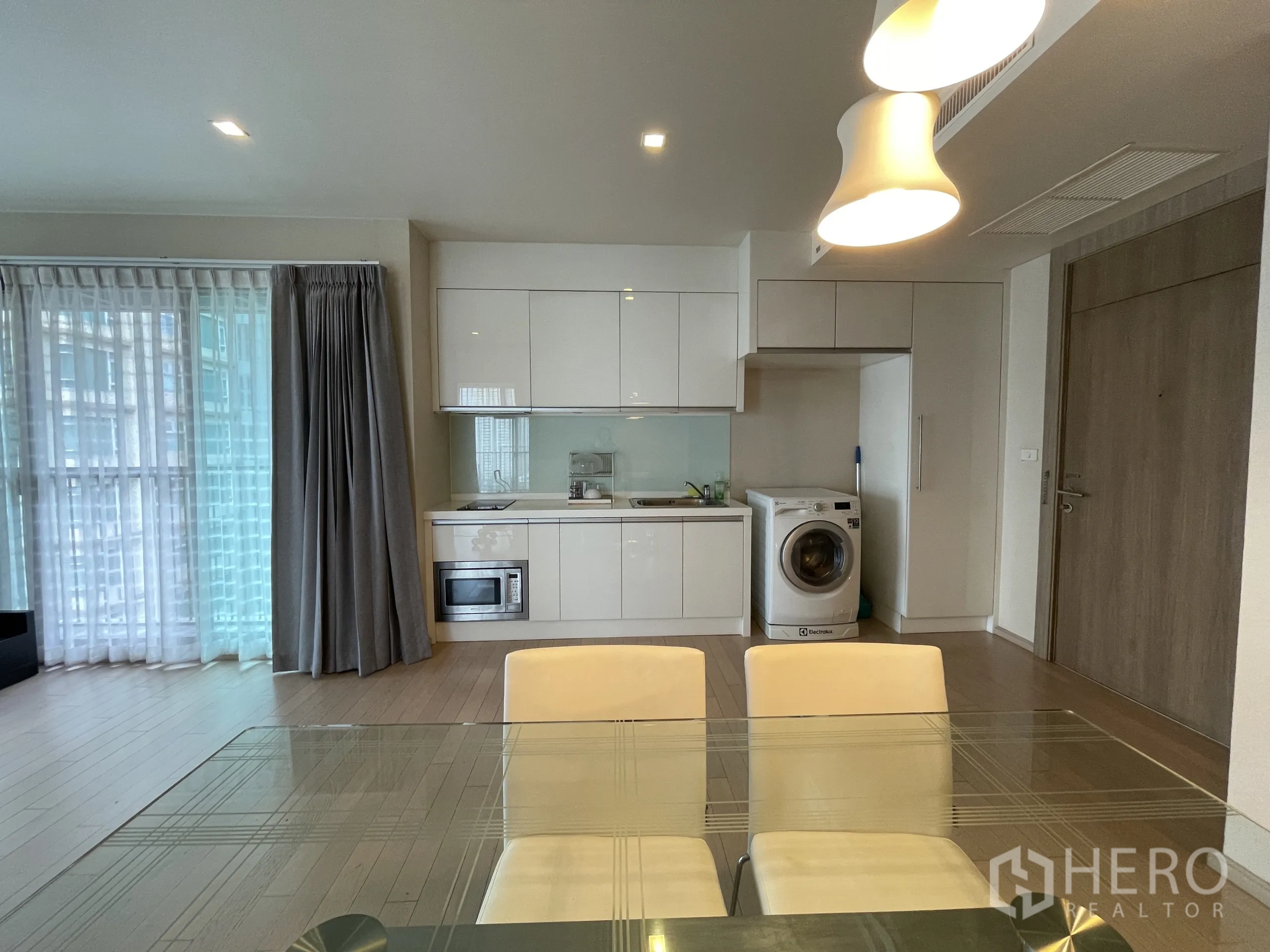 公寓 出租在Watthana, Bangkok - 从餐桌望向厨房，亮面橱柜、水槽、微波炉、洗衣机与落地窗帘。